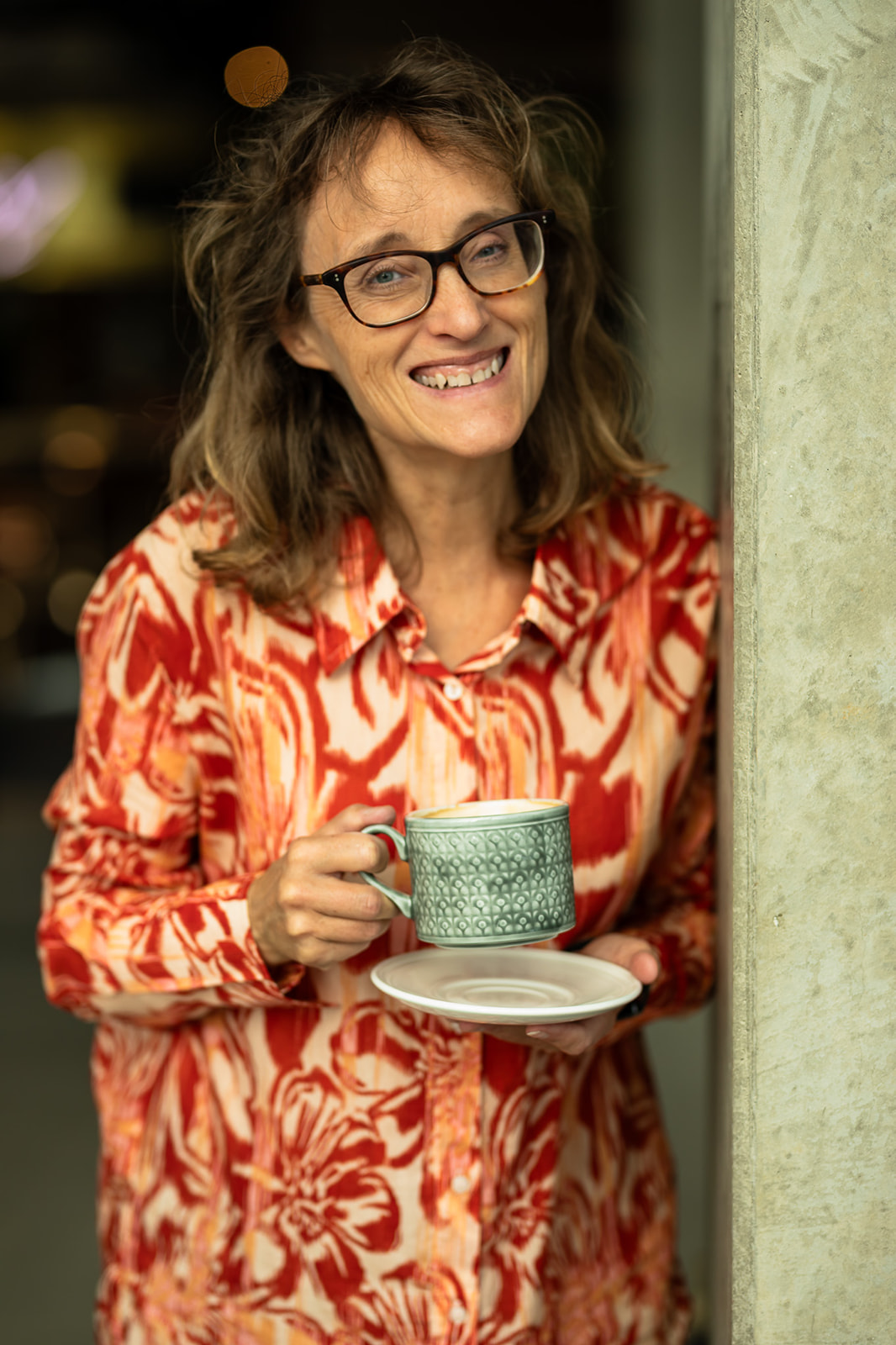 Anna står skrattande lutad mot en stople med en kaffekopp med fat i handen. Mönstrad röd skjorta