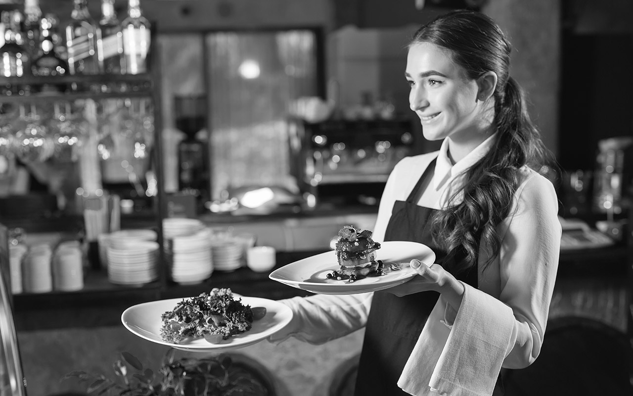 En ung kvinna med mörkt hår uppsatt i en låg hästsvans bär en tallrik i vardera hand i restaurangmiljö. Visar servering